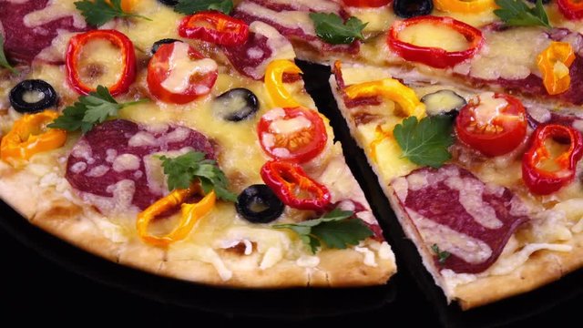 Homemade hot pizza with salami and vegetables just from oven with cut off piece rotates on black background in 4K. Traditional tasty food.
