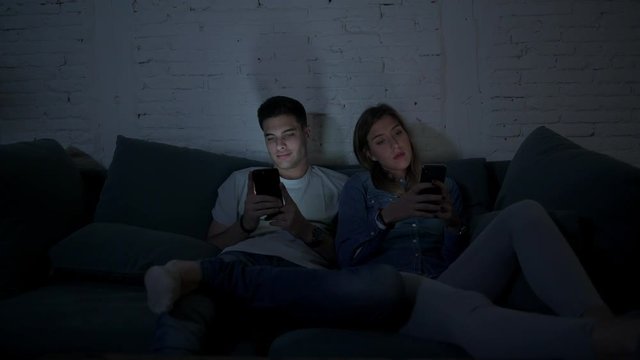 young couple on their 20s using mobile phone ignoring each other sitting at home couch concentrated on internet app in relationship lack of communication and smart phone addiction concept