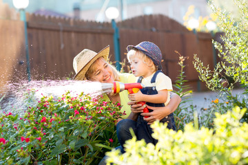 Pretty woman and her child son watering flowers with water hose in the yard