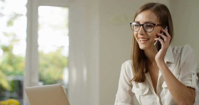 4K Busy professional woman talking on the phone while working on laptop at home