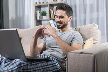 Portrait of cheerful male sitting on sofa with laptop, he is looking at screen and making heart figure with hands