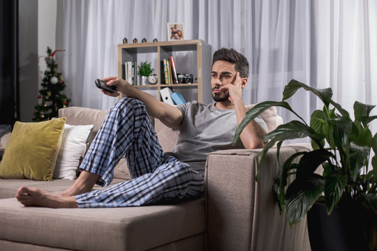 Sad young male relaxing on comfortable couch with tv remote. Big houseplant standing beside him