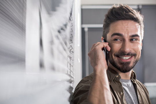 Portrait of contented handsome man in office chatting by mobile phone. Copy space in left side