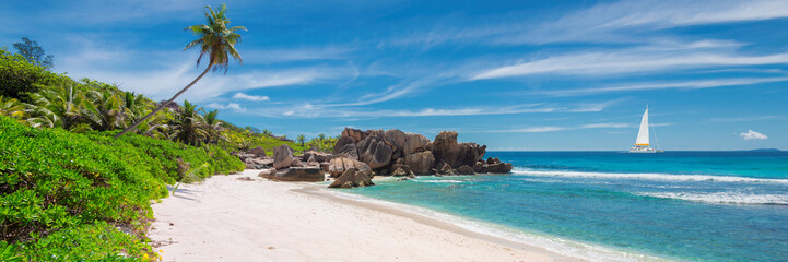 Sailing boat in the turquoise sea and sandy beach with palm trees and beautiful rocks on Seychelles.