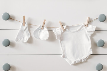 Set of baby clothes for newborn in white color. Baby socks, mittens and bodysuit are hanging on a...