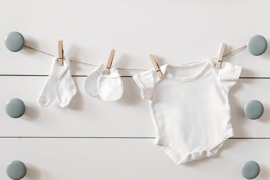 Set of baby clothes for newborn in white color. Baby socks, mittens and bodysuit are hanging on a clothline. Newborn concept background.