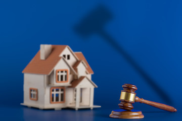 Gavel and model house on blue background. The gavel casts an ominous shadow over the house. Concept for house auction or real estate law