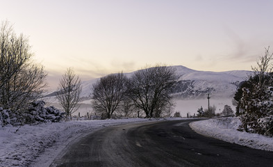 Fototapeta na wymiar A road in the Scottish countryside in the Highlands surrounded by snow with mountains and a beautiful pink sunset in the background with clear sky ideal for copyspace