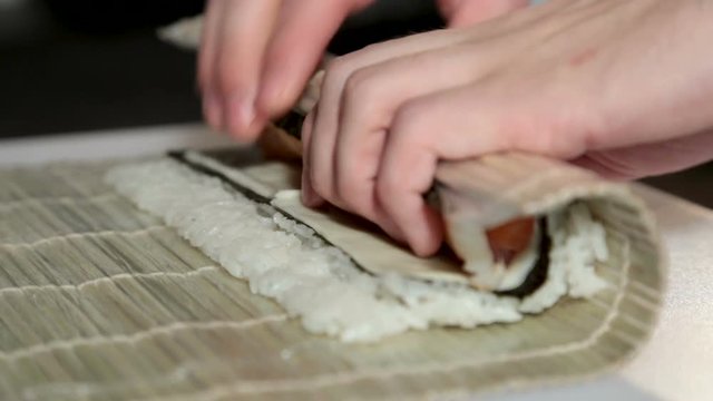 A man with his bare hands wraps a sushi with a shrimp and salmon. On the table is a sheet of nori, a bamboo mat, rice, salmon and shrimp. Close-up