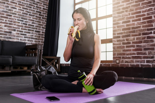 Portrait of young caucasian woman resting after training holding a sports bottle sitting on mat with legs crossed in gym with loft interior