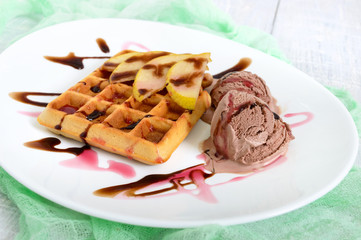 Crispy Belgian waffle with pieces of pear, berry and chocolate syrup and ice cream balls on a white plate.
