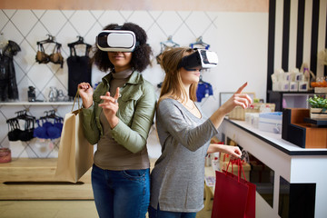 Two women in modern virtual reality headsets having expirience in shopping at lingerie store. Multiracial girls in vr glasses with bags touching and pointing interface elements in underwear shop.