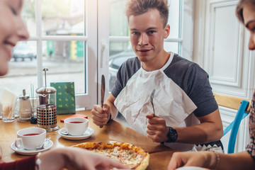 Stylish teenager wearing napkin holding knife and fork ready to eat pizza sitting against window at restaurant