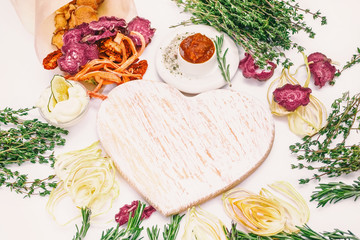 Homemade colorful chips of different fresh vegetables - beets, sweet potatoes, carrots, cucumber, onions, rosemary and thyme on a light background around the white Board in the shape of a heart.