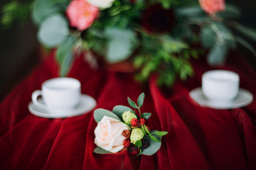 close-up of a boutonnier with a pink rose which lies on a table with a red tablecloth on the background of cups and flowers