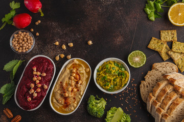 Vegan food background. Vegetarian snacks: hummus, beetroot hummus, green peas dip, vegetables. Top view, dark background, copy space.