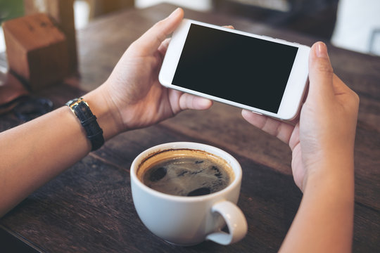 Mockup image of hands holding white mobile phone with blank black screen for watching and playing games with a cup of coffee on wooden table in cafe