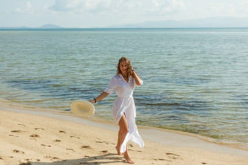 smiling girl in white dress with hat walking on tropical beach at the sea