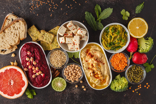Vegan food background. Vegetarian snacks: hummus, beetroot hummus, green peas dip, vegetables, cereals, tofu. Top view, dark background, copy space.