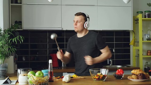 Attractive young funny man dancing and singing with ladle while cooking in the kitchen at home