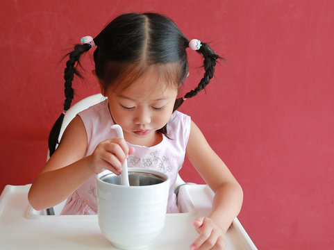 Asian child girl eating pork bone soup on high chair against red wall background at restaurant.