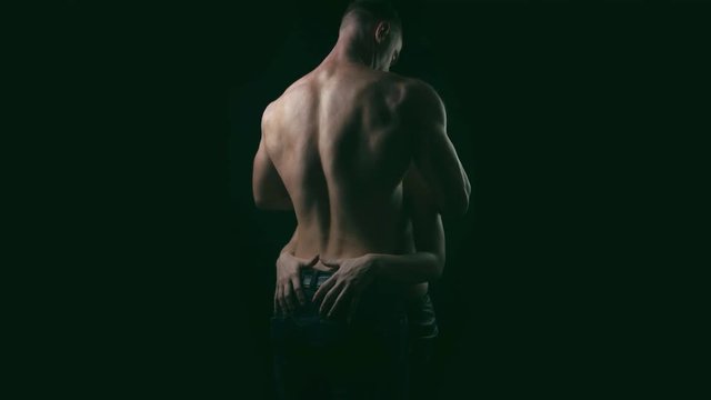 Blond woman slowly touching back of young muscular shirtless man on black background. 