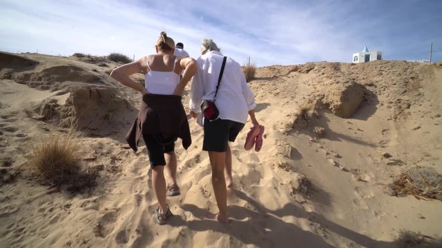 Following three generations as they climb a steep sandy trail with blue sky above.