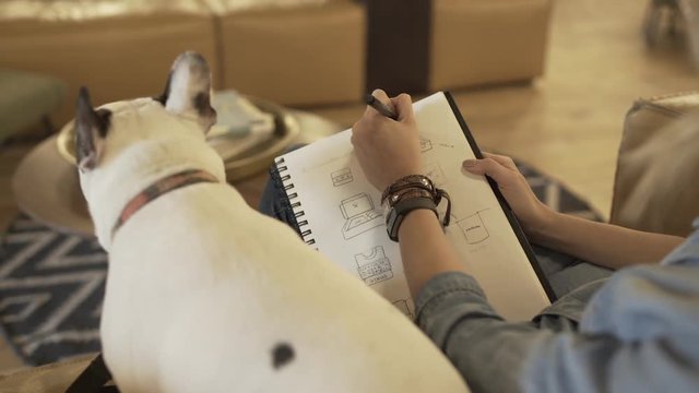 A girl sits on a yellow couch with her dog, brainstorming and drawing logo concepts on a notebook.
