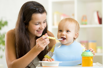 Mommy giving healthy food to her baby son on high chair in kitchen.