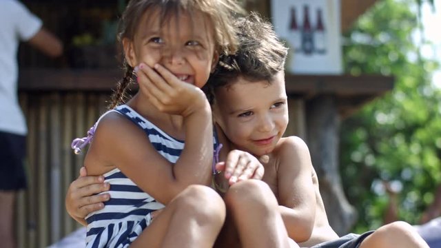 Two preschool children sitting together at the beach, smiling and hugging each other, their hair blowing in the wind
