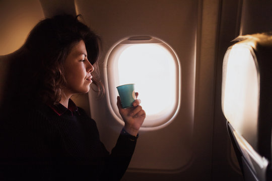 Young Caucasian  Woman Drinking Coffee  Inside Airplane Sitting On Passenger Seat Near Window, Relaxing  And Dreaming About Vacations.