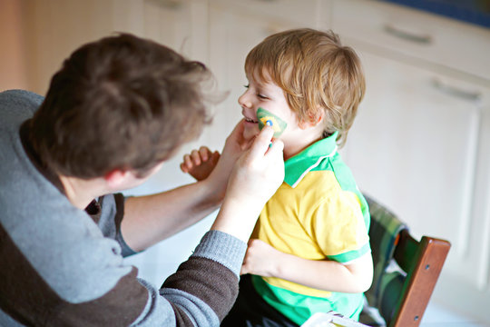 Young Dad Painting Flag On Face Of Little Son For Football Or Soccer Game.