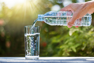 Hand, die Trinkwasserflasche hält, die Wasser in Glas auf Holztisch auf unscharfem grünem Naturhintergrund gießt