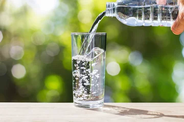 Foto op Plexiglas Hand met drinkwaterfles die water in glas giet op houten tafelblad op vage groene bokehachtergrond met zacht zonlicht © shark749