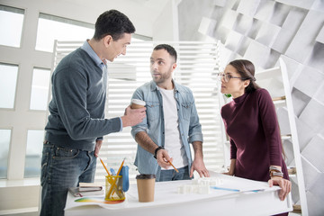Think tank. Good-looking concentrated young bearded man holding a pencil and standing at the table with his colleagues and discussing work with them