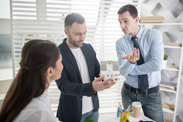 Future house. Handsome alert young dark-haired man smiling and holding a house miniature while his colleagues standing near him