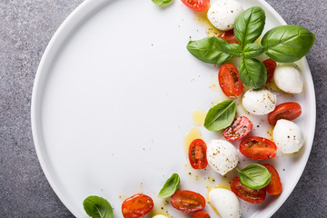 White plate of healthy classic delicious caprese salad with ripe tomatoes and mozzarella cheese with fresh basil leaves on gray concrete background with space for text. Italian food. Top view.