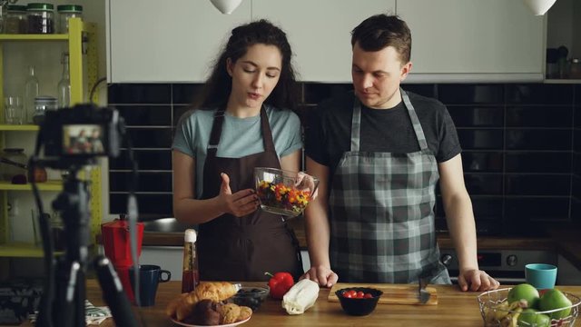 Smiling attractive couple recording video food vlog about cooking haelthy salad on digital camera in the kitchen at home. Vlogging, education and social media concept