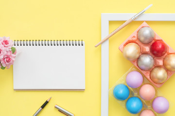 Flat lay top view colorful easter egg painted in pastel colors composition and mock up blank notebook with spring flowers on yellow pastel background. Easter day background top view with copy space.
