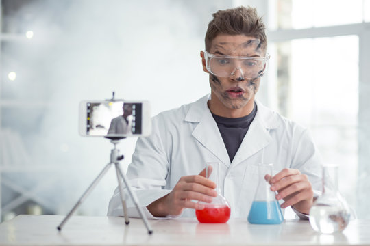 Surprised. Handsome Perplexed Fair-haired Young Blogger Wearing Uniform And Holding Test Tubes And Making New Video