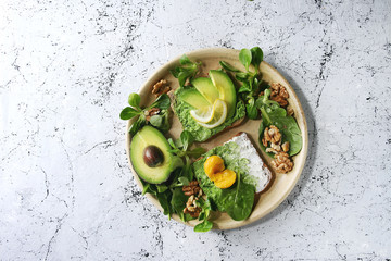 Vegetarian sandwiches with avocado, ricotta, egg yolk, spinach, walnuts on whole grain toast bread on ceramic plate with ingredients above over white marble background. Top view, space
