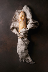 Loaf of sliced fresh baked artisan baguette bread wrapped in linen cloth over dark brown texture background. Top view, copy space.