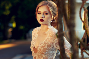 Naklejka premium Blonde woman with elegant hairstyle wearing dress and white lace jacket, standing on garden terrace and leaning on balustrade decorated with large flower vases against shining sun on background.