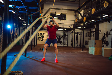 Man with battle ropes exercise in the fitness gym. Young male wearing sportswear.