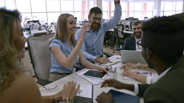 Group of six colleagues sitting at table in office and giving high five to each other after successful work on project, camera moving forwards