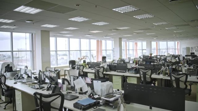 Dolly shot of big empty office with large windows and many laptop computers and papers on tables, shot in daytime, camera moving forwards