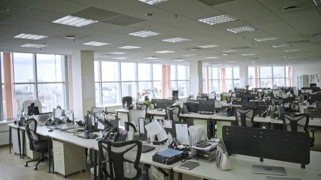Dolly shot of big empty office with large windows and many laptop computers and papers on tables, shot in daytime, camera moving backwards