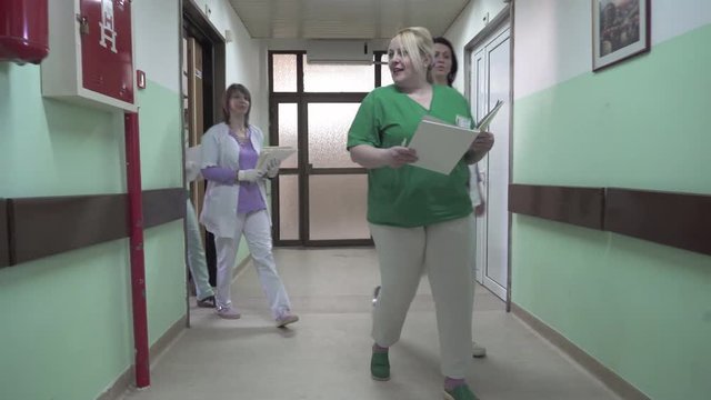 Team of nurses with medical documentation passing through the hallway of hospital ward and visiting senior male patient who lying in bed, female medic technician pushing wheelchair, gimbal shot