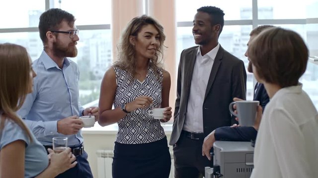 Group of six colleagues standing in semicircle and having friendly conversation over cup of tea, tilt up shot