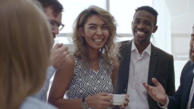 Group of colleagues standing together, talking and laughing over cup of tea during break from work
